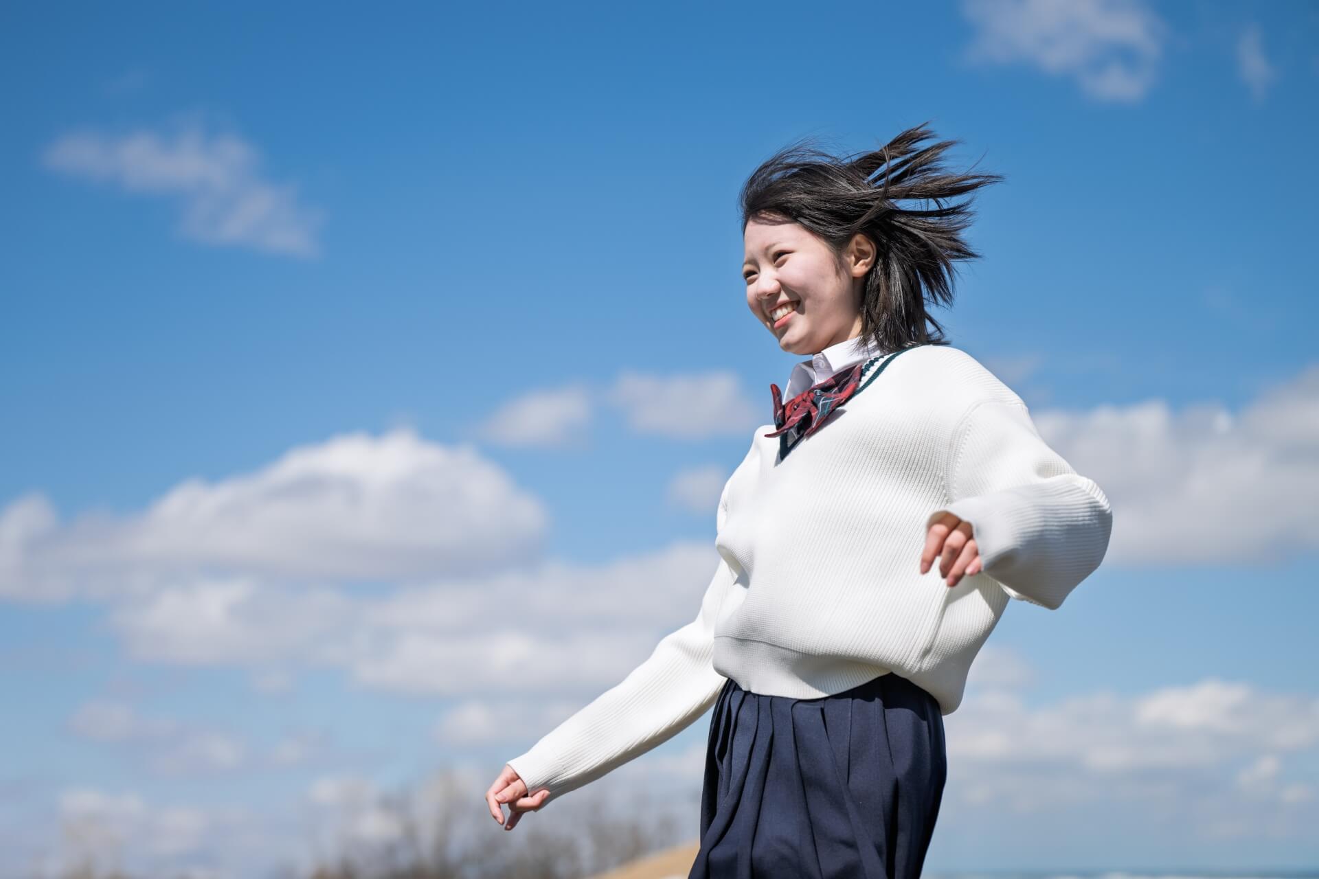 青空を背景に、風に髪をなびかせながら微笑む制服姿の女子高生