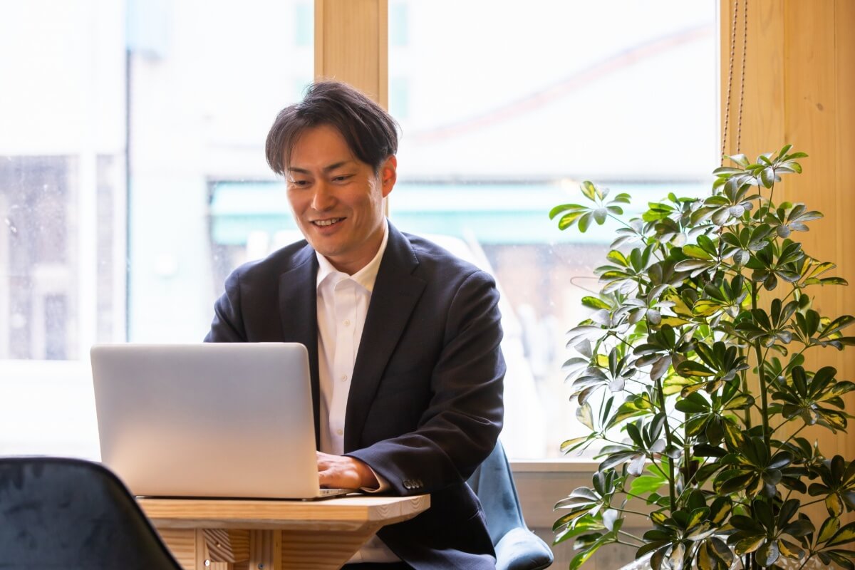 窓際の席でノートパソコンを使って作業をするスーツ姿のビジネスマン。観葉植物が横に置かれている