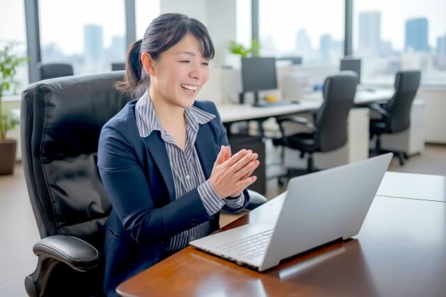 オフィスのデスクでノートパソコンを前に拍手をする笑顔のビジネスウーマン。都市の景色が見える窓を背景にしている