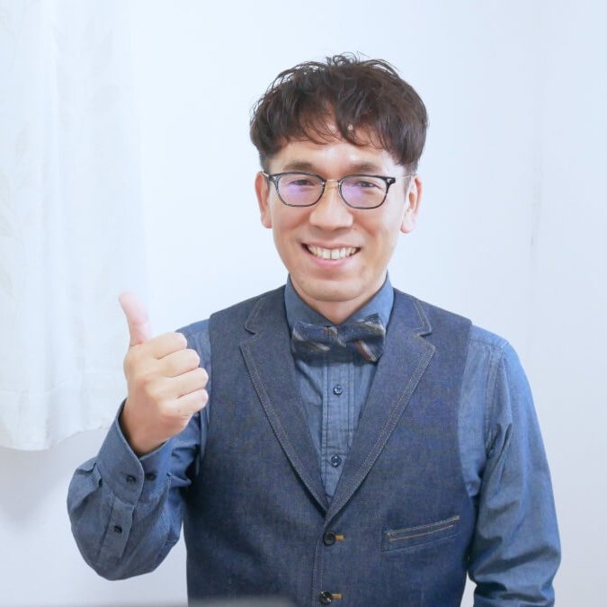 ベストと蝶ネクタイを着用し、笑顔でこちらを見る代表理事カクモト・ジン