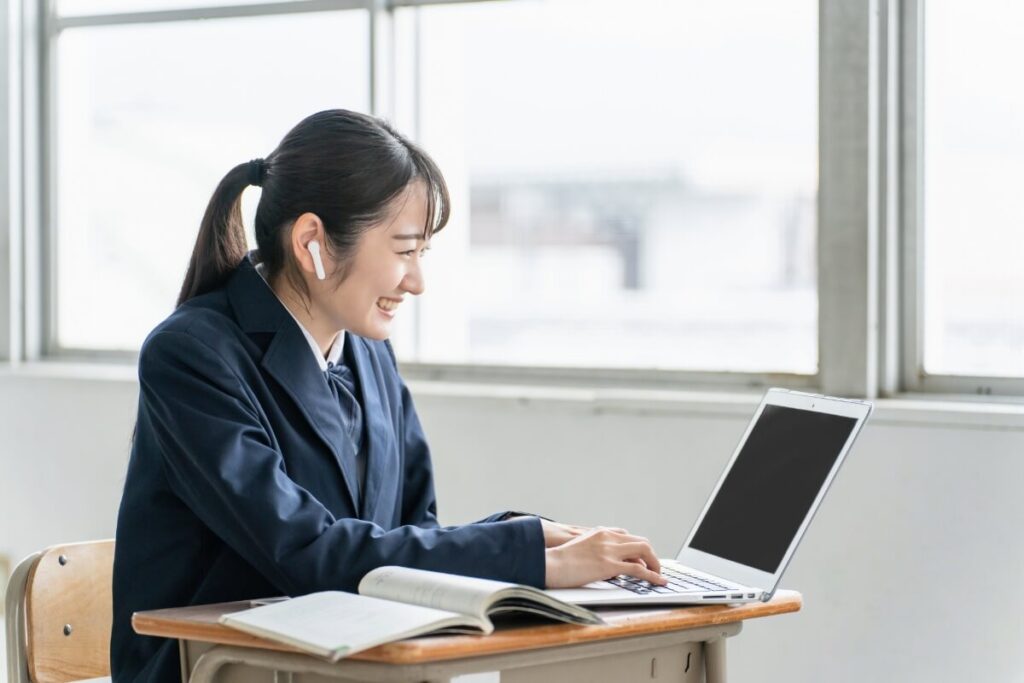 ワイヤレスイヤホンを着けてノートパソコンで学習する笑顔の女子学生