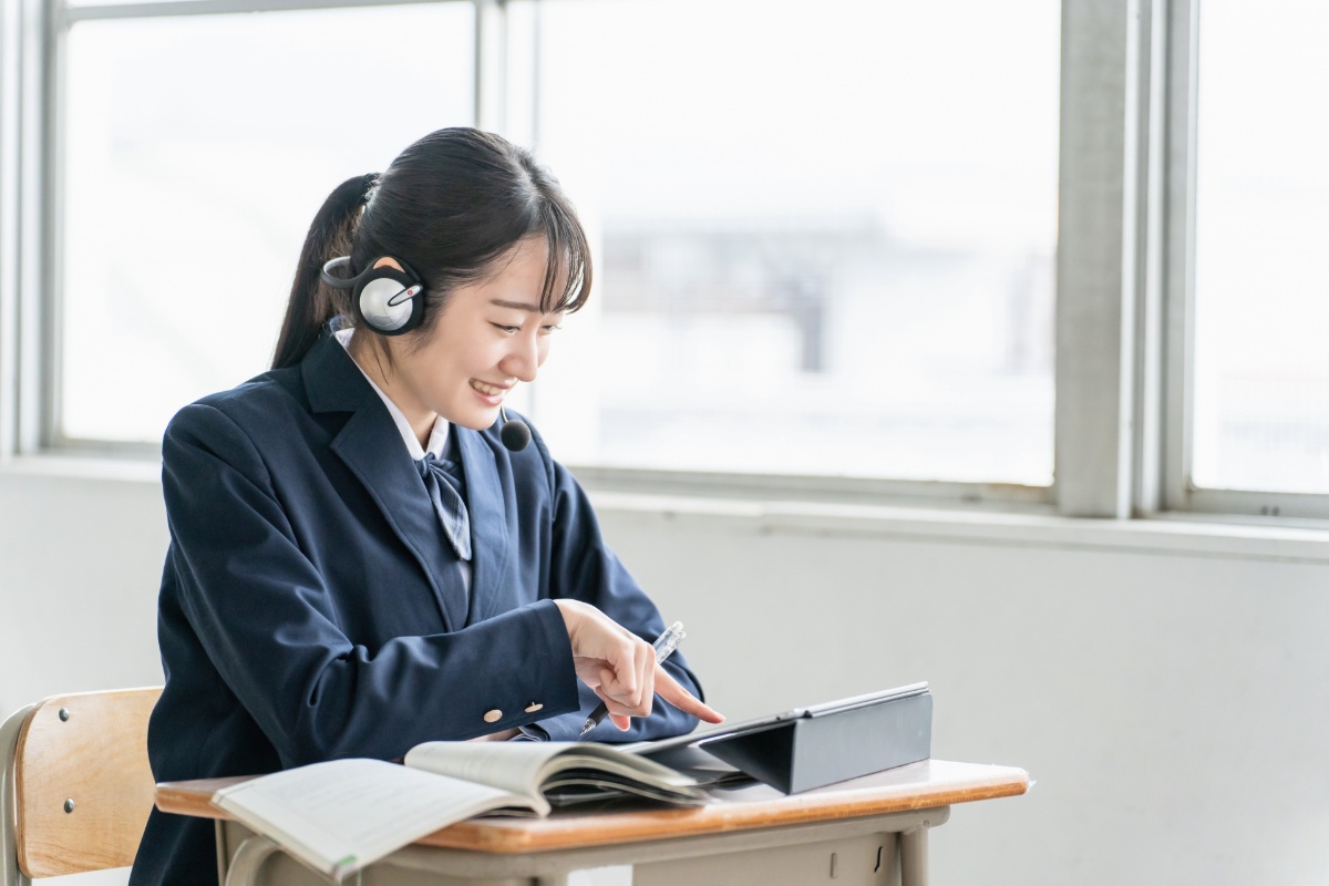 教室でヘッドセットを着けてタブレット端末と教科書を使いながらオンライン学習をする制服姿の女子学生
