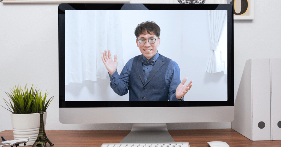 デスクトップモニターに映る笑顔で手を振る眼鏡をかけた男性とのビデオ通話。木製デスクの上には観葉植物とキーボードが置かれている