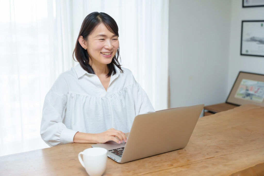 自宅の明るいデスクでコーヒーカップを置いてノートパソコンで作業をする笑顔の女性